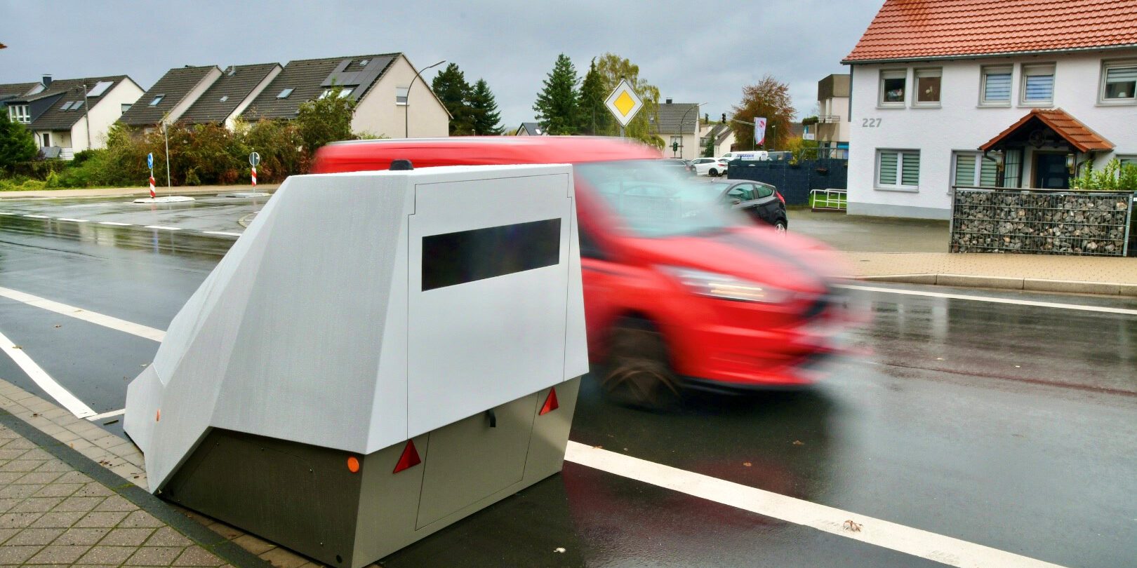 Mobiler Blitzer am Straßenrand in einem Wohngebiet; ein rotes Auto fährt mit Bewegungsunschärfe vorbei, die Straße ist nass vom Regen.