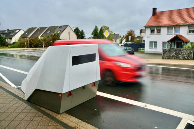 Foto: Ralf Engel Mobiler Blitzer am Straßenrand in einem Wohngebiet; ein rotes Auto fährt mit Bewegungsunschärfe vorbei, die Straße ist nass vom Regen.