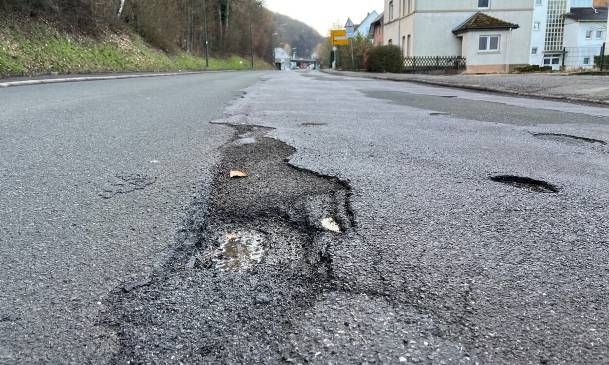 Eine Straße mit mehreren Schlaglöchern und Rissen im Asphalt, im Vordergrund ist ein ausgefahrenes Loch mit Wasser zu sehen. Am Straßenrand stehen Häuser, Bäume und Verkehrsschilder.