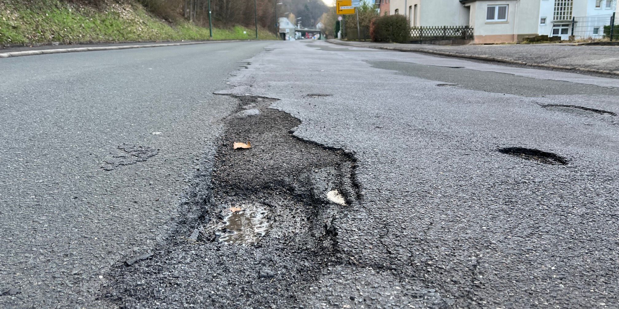 Eine Straße mit mehreren Schlaglöchern und Rissen im Asphalt, im Vordergrund ist ein ausgefahrenes Loch mit Wasser zu sehen. Am Straßenrand stehen Häuser, Bäume und Verkehrsschilder.