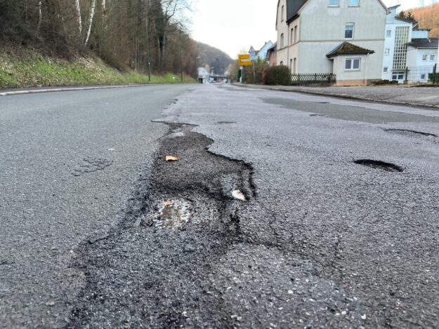 Eine Straße mit mehreren Schlaglöchern und Rissen im Asphalt, im Vordergrund ist ein ausgefahrenes Loch mit Wasser zu sehen. Am Straßenrand stehen Häuser, Bäume und Verkehrsschilder.