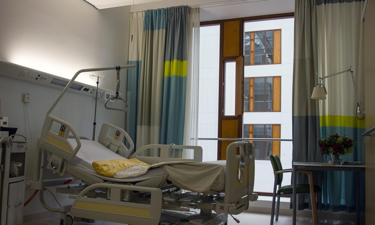 Leeres Patientenzimmer in einem Krankenhaus mit verstellbarem Pflegebett, Infusionsständer und Vorhängen vor einem großen Fenster; daneben ein kleiner Tisch mit Stuhl und Blumenstrauß.