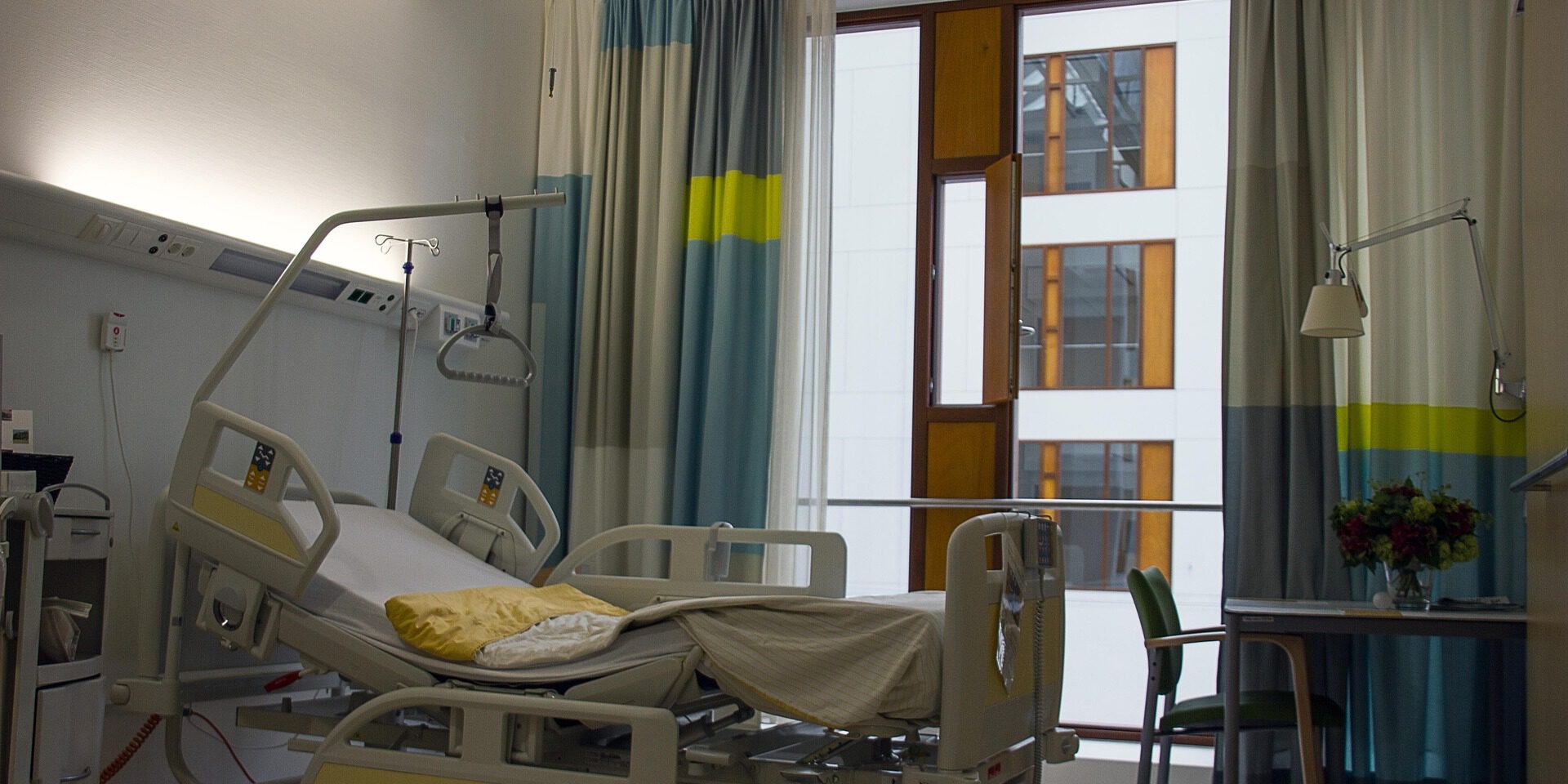 Leeres Patientenzimmer in einem Krankenhaus mit verstellbarem Pflegebett, Infusionsständer und Vorhängen vor einem großen Fenster; daneben ein kleiner Tisch mit Stuhl und Blumenstrauß.