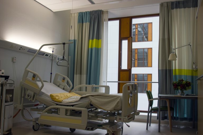 Leeres Patientenzimmer in einem Krankenhaus mit verstellbarem Pflegebett, Infusionsständer und Vorhängen vor einem großen Fenster; daneben ein kleiner Tisch mit Stuhl und Blumenstrauß.