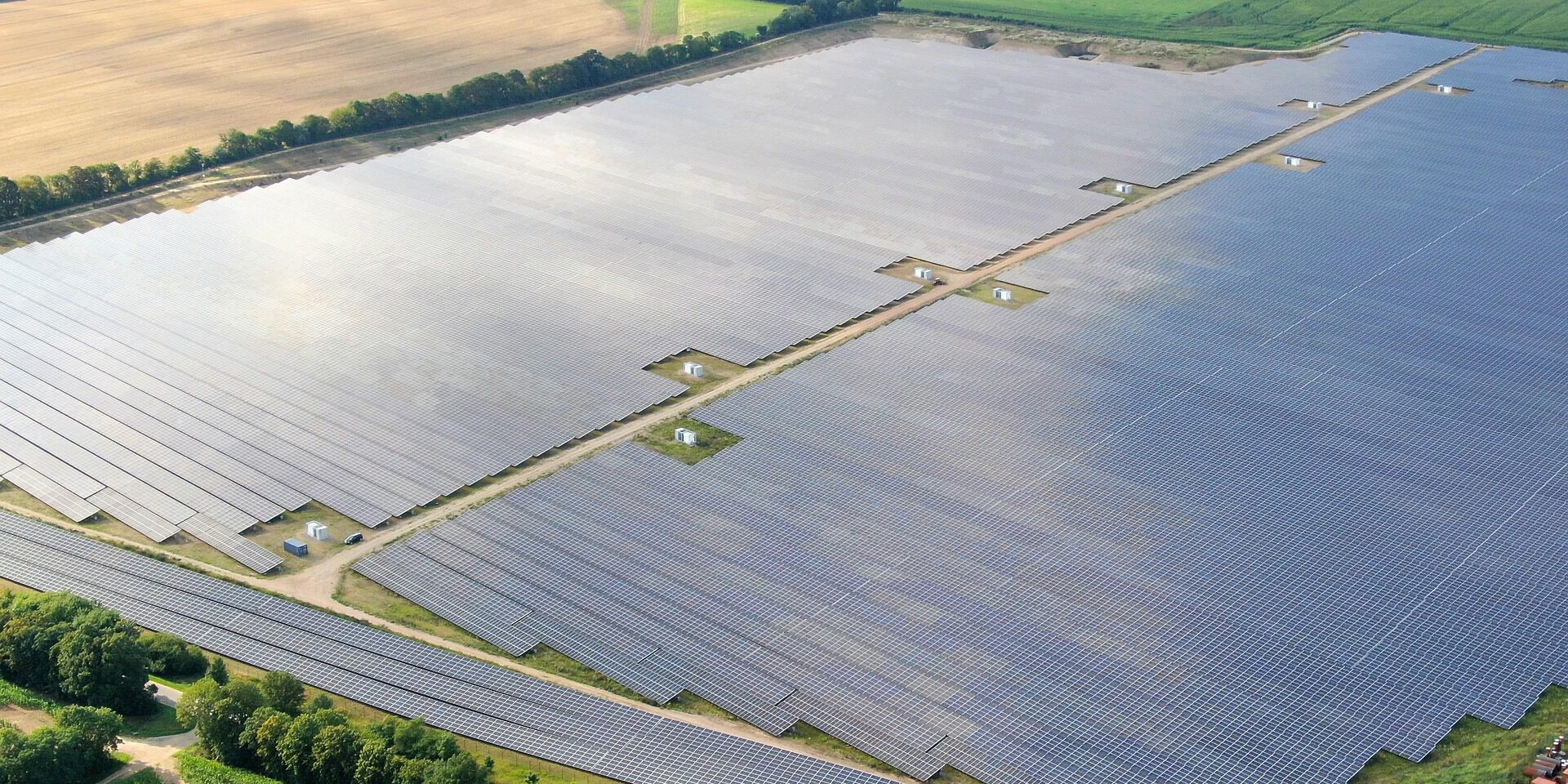 Eine große Photovoltaikanlage erstreckt sich über weite Flächen in einer ländlichen Umgebung. Reihen aus Solarmodulen liegen zwischen Feldern und Baumreihen und sind aus der Luft aufgenommen.