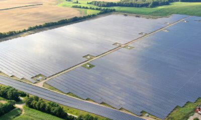 Eine große Photovoltaikanlage erstreckt sich über weite Flächen in einer ländlichen Umgebung. Reihen aus Solarmodulen liegen zwischen Feldern und Baumreihen und sind aus der Luft aufgenommen.