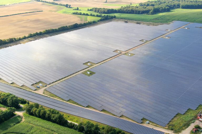 Eine große Photovoltaikanlage erstreckt sich über weite Flächen in einer ländlichen Umgebung. Reihen aus Solarmodulen liegen zwischen Feldern und Baumreihen und sind aus der Luft aufgenommen.