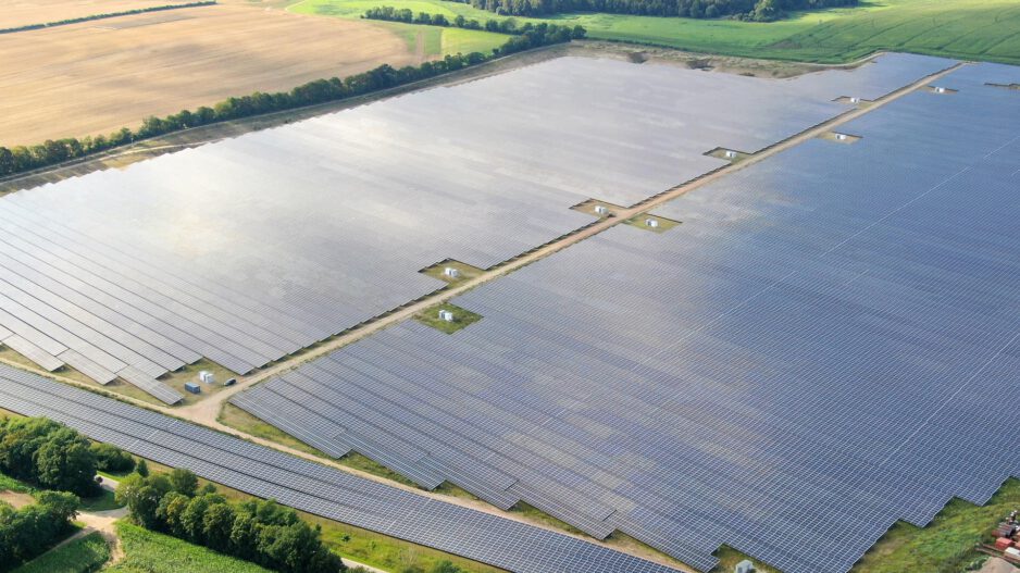 Eine große Photovoltaikanlage erstreckt sich über weite Flächen in einer ländlichen Umgebung. Reihen aus Solarmodulen liegen zwischen Feldern und Baumreihen und sind aus der Luft aufgenommen.