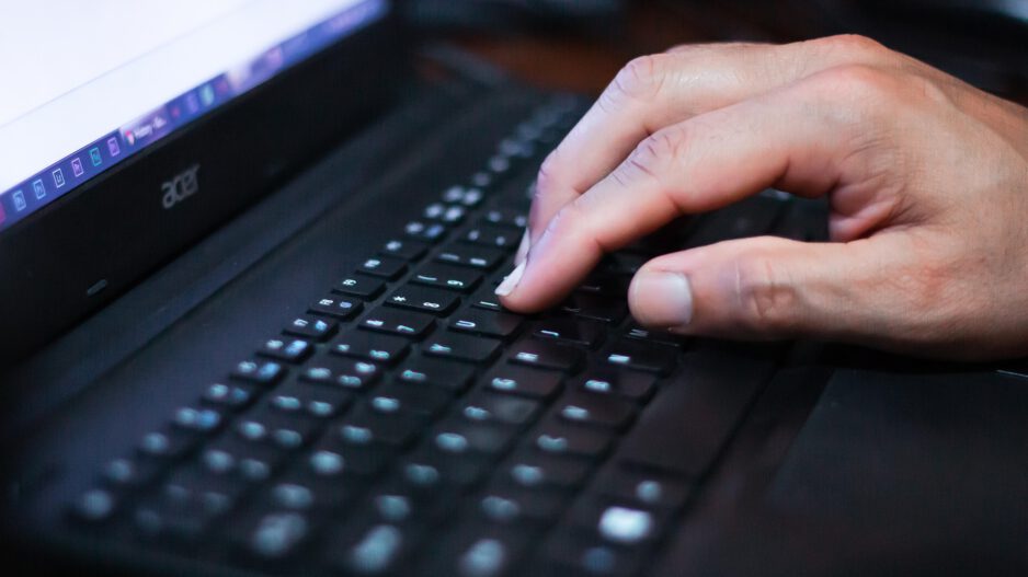 Eine Hand tippt auf der Tastatur eines Laptops, der Bildschirm ist unscharf im Hintergrund zu sehen.