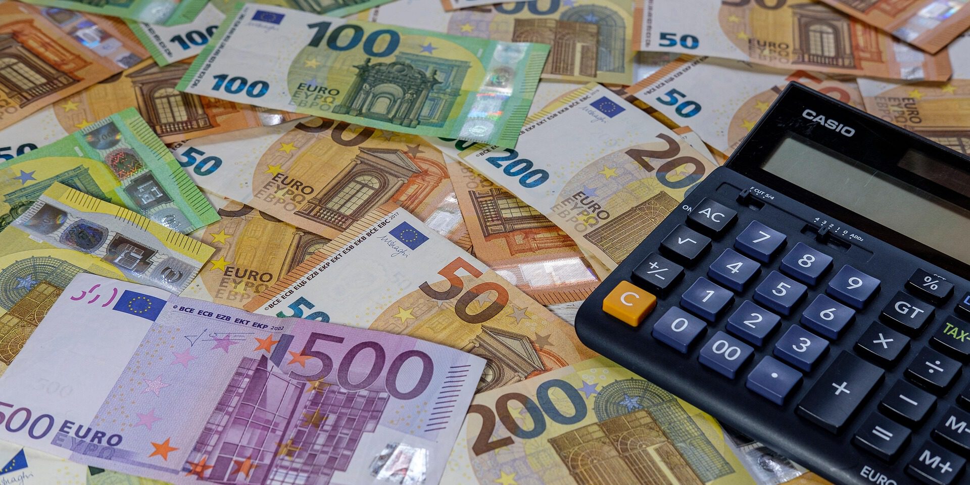 Viele Euro-Banknoten verschiedener Stückelungen liegen ausgebreitet auf einem Tisch, daneben befindet sich ein Taschenrechner.