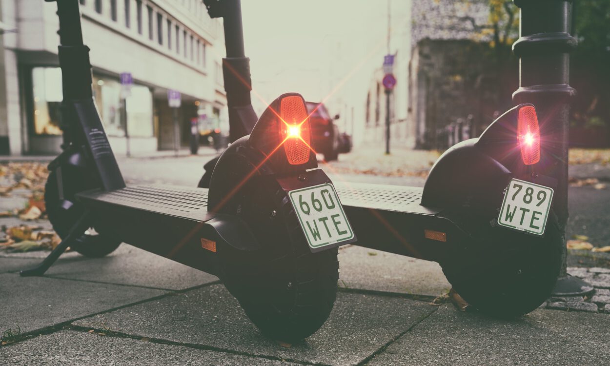 Zwei geparkte E-Scooter stehen nebeneinander auf einem Gehweg in einer Stadtstraße. Die roten Rücklichter leuchten, an den Hinterrädern sind Kennzeichen angebracht, im Hintergrund sind Gebäude und parkende Autos unscharf zu erkennen.