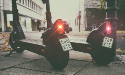 Zwei geparkte E-Scooter stehen nebeneinander auf einem Gehweg in einer Stadtstraße. Die roten Rücklichter leuchten, an den Hinterrädern sind Kennzeichen angebracht, im Hintergrund sind Gebäude und parkende Autos unscharf zu erkennen.