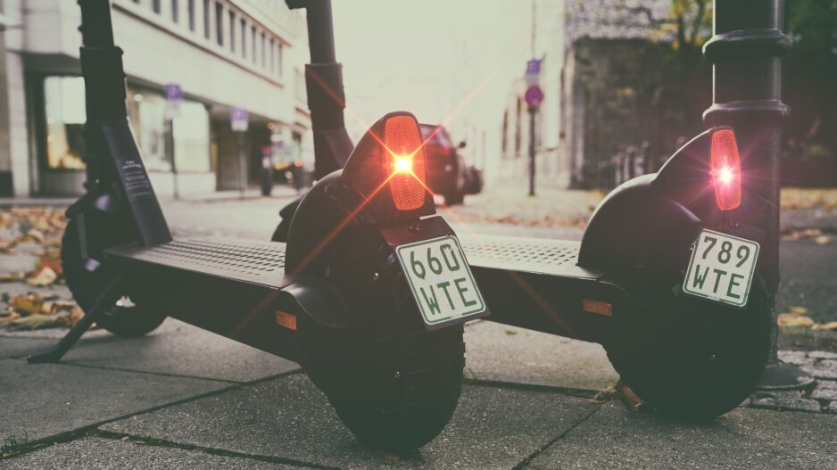 Zwei geparkte E-Scooter stehen nebeneinander auf einem Gehweg in einer Stadtstraße. Die roten Rücklichter leuchten, an den Hinterrädern sind Kennzeichen angebracht, im Hintergrund sind Gebäude und parkende Autos unscharf zu erkennen.