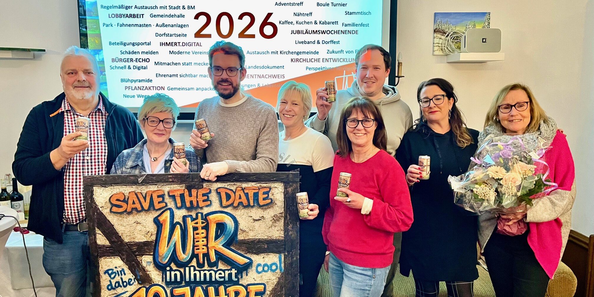 Acht Personen stehen vor einer Präsentationsfolie mit der Jahreszahl 2026 und halten Getränkedosen; davor ein Schild mit „Save the Date – Wir in Ihmert, 10 Jahre“.