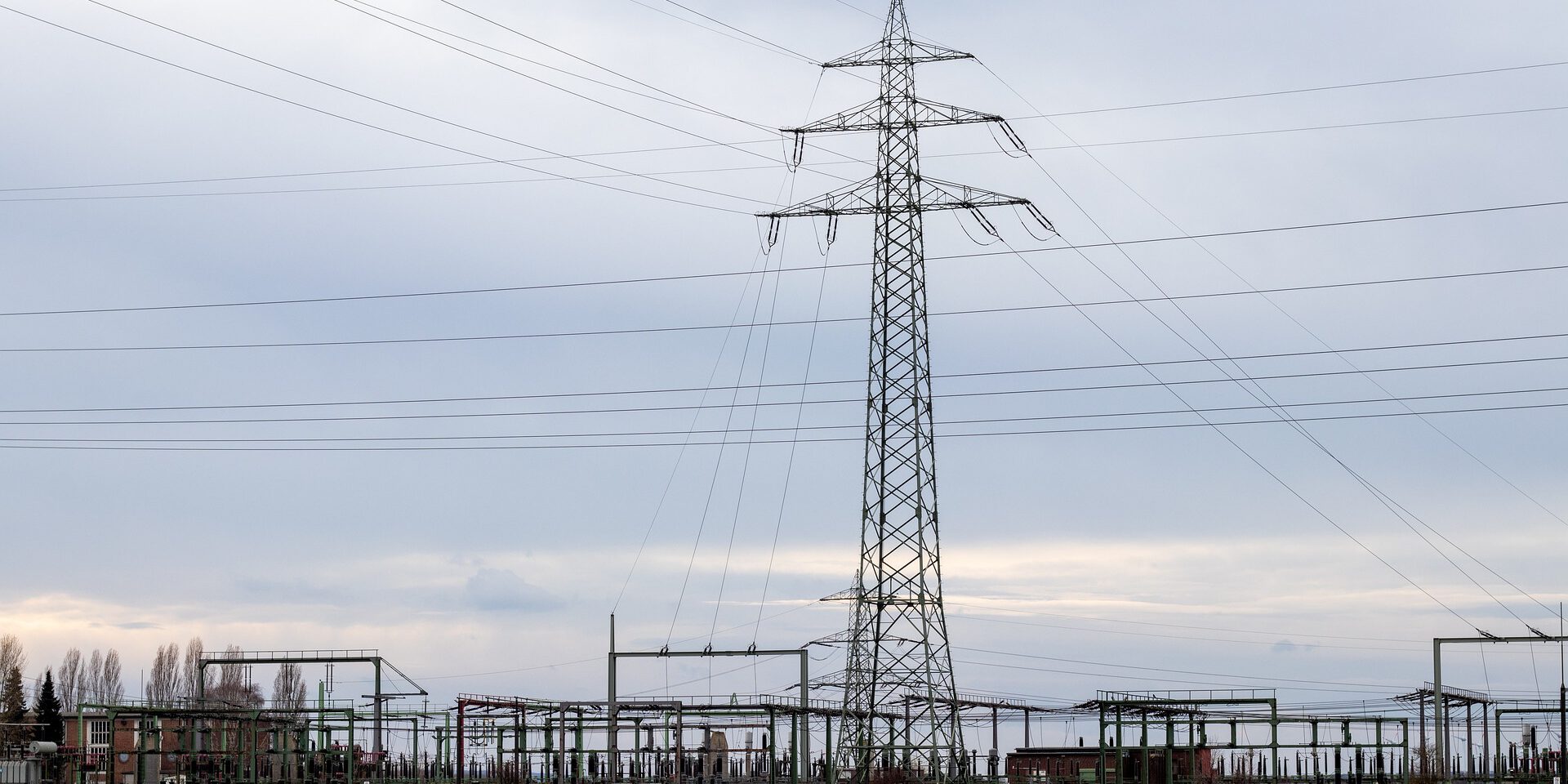 Umspannwerk mit Stromleitungen und hohem Hochspannungsmast in einer offenen Landschaft unter bewölktem Himmel.
