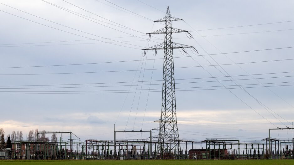 Umspannwerk mit Stromleitungen und hohem Hochspannungsmast in einer offenen Landschaft unter bewölktem Himmel.