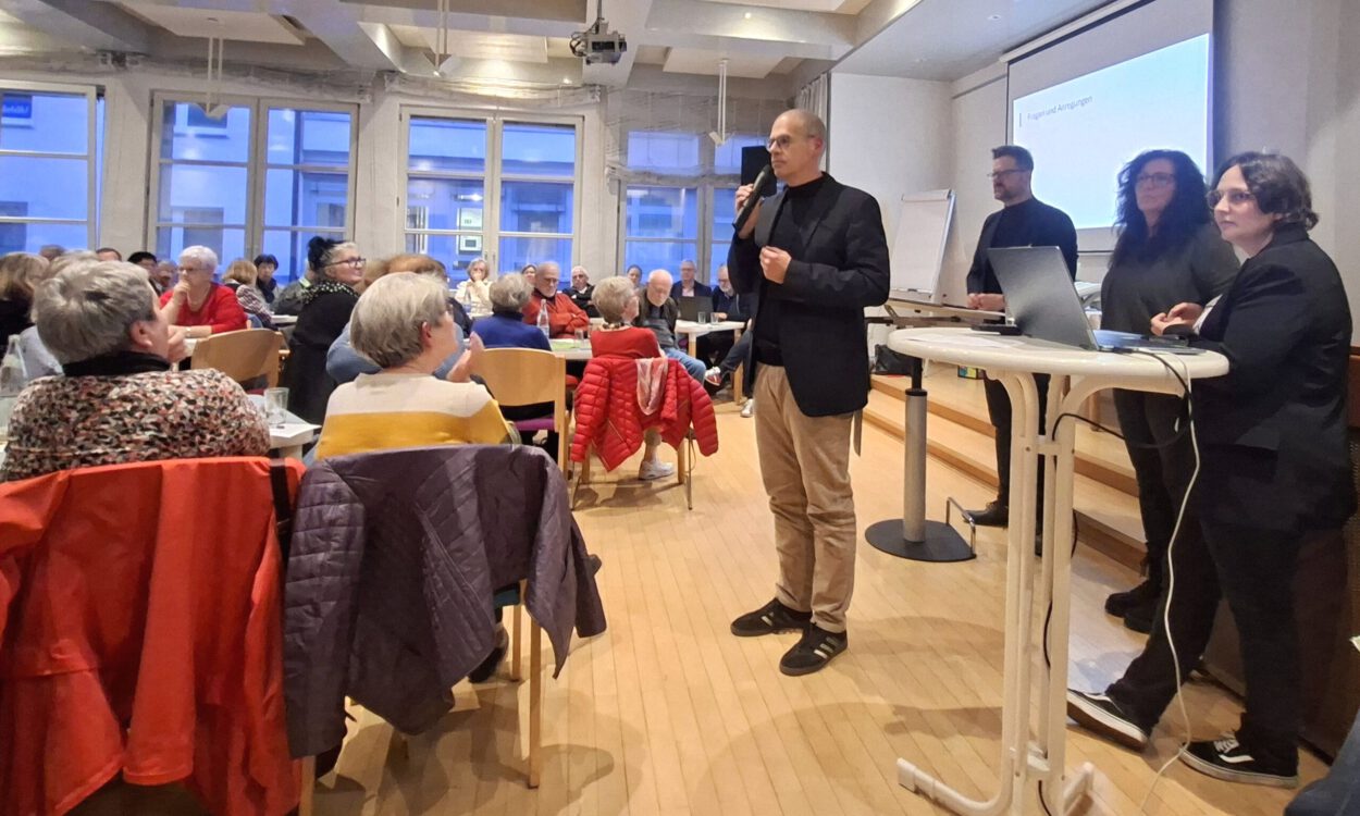 Ein Mann spricht mit Mikrofon vor Publikum in einem Veranstaltungsraum, während mehrere Personen neben ihm an einem Tisch stehen.