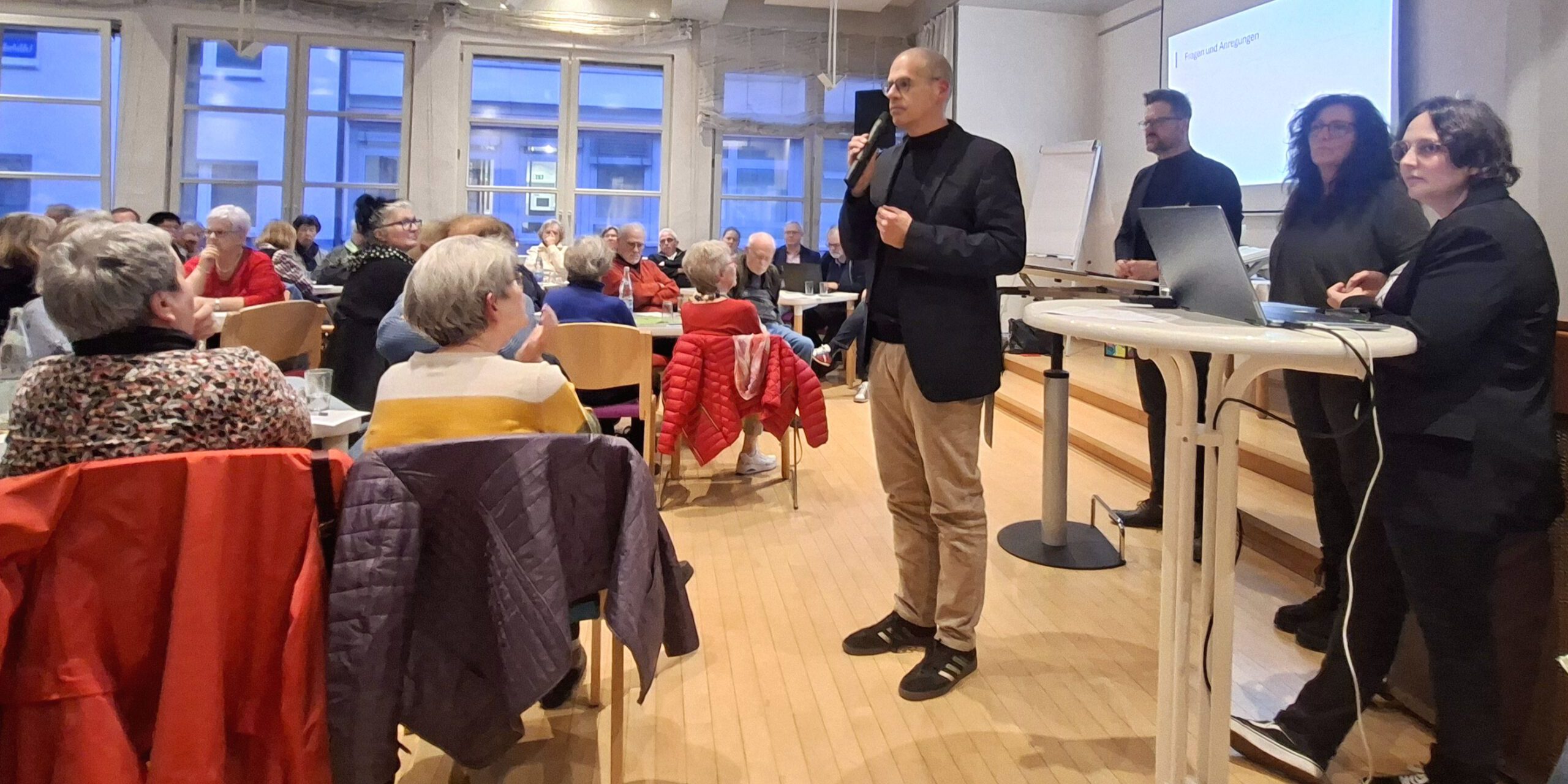 Ein Mann spricht mit Mikrofon vor Publikum in einem Veranstaltungsraum, während mehrere Personen neben ihm an einem Tisch stehen.