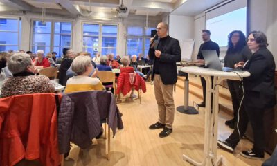 Ein Mann spricht mit Mikrofon vor Publikum in einem Veranstaltungsraum, während mehrere Personen neben ihm an einem Tisch stehen.