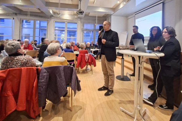 Ein Mann spricht mit Mikrofon vor Publikum in einem Veranstaltungsraum, während mehrere Personen neben ihm an einem Tisch stehen.