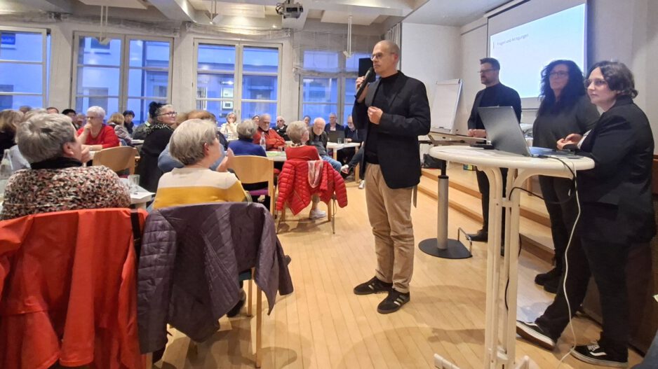 Ein Mann spricht mit Mikrofon vor Publikum in einem Veranstaltungsraum, während mehrere Personen neben ihm an einem Tisch stehen.