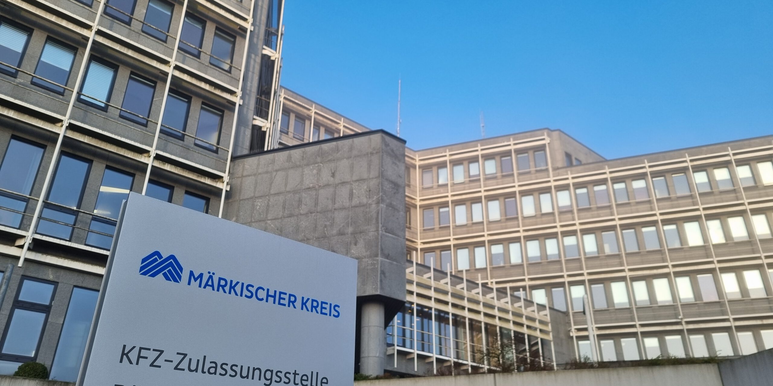 Schild mit der Aufschrift „Märkischer Kreis – KFZ-Zulassungsstelle, Bürgerbüro“ vor einem mehrstöckigen Verwaltungsgebäude.