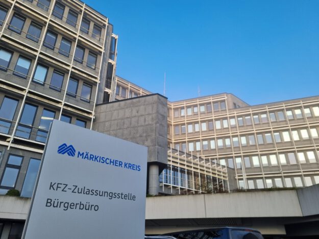 Schild mit der Aufschrift „Märkischer Kreis – KFZ-Zulassungsstelle, Bürgerbüro“ vor einem mehrstöckigen Verwaltungsgebäude.