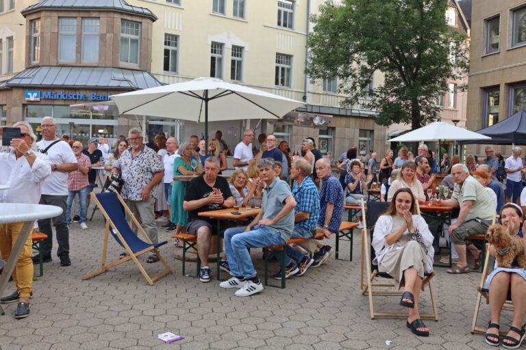 Viele Menschen sitzen und stehen bei einer Veranstaltung auf einem Platz vor der Märkischen Bank und schauen in eine Richtung.