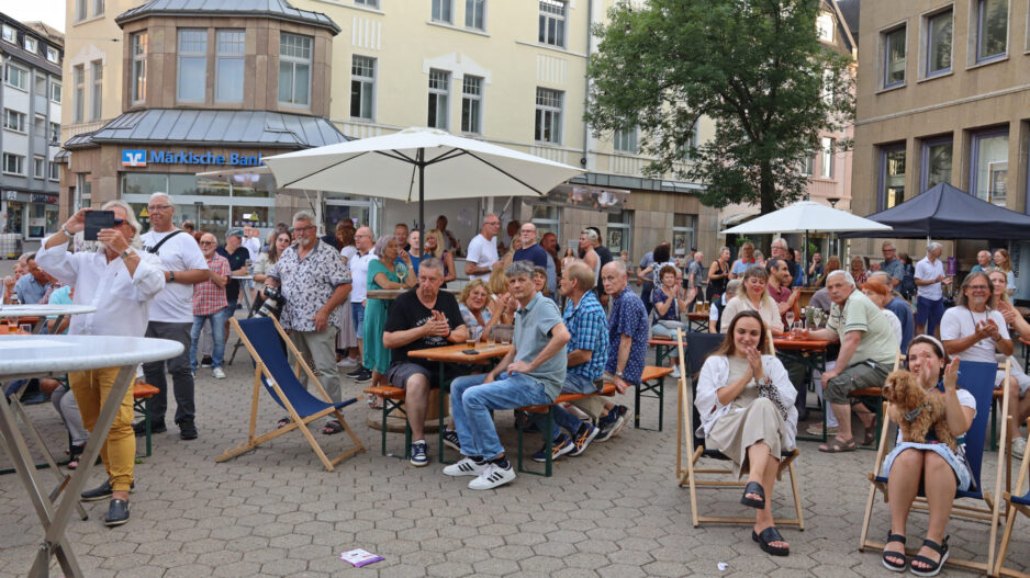 Viele Menschen sitzen und stehen bei einer Veranstaltung auf einem Platz vor der Märkischen Bank und schauen in eine Richtung.