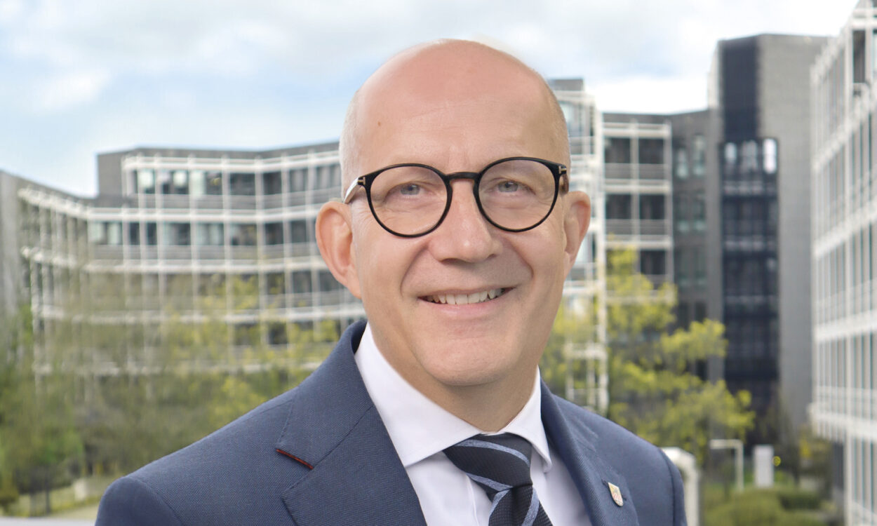 Ein Mann mit Glatze und runder Brille im Anzug lächelt vor modernen Bürogebäuden im Hintergrund.