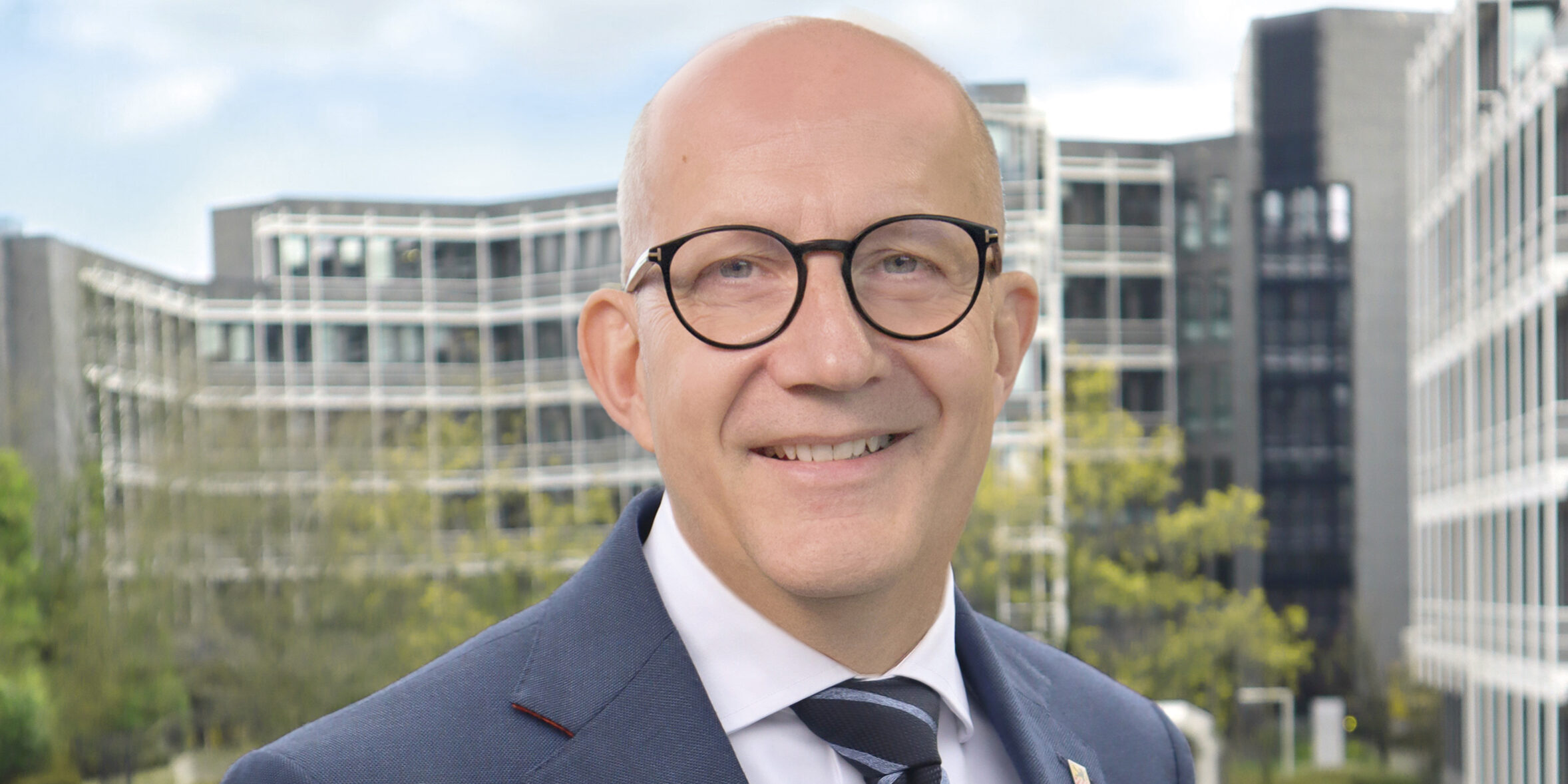 Ein Mann mit Glatze und runder Brille im Anzug lächelt vor modernen Bürogebäuden im Hintergrund.