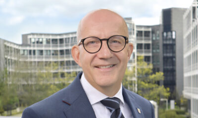 Ein Mann mit Glatze und runder Brille im Anzug lächelt vor modernen Bürogebäuden im Hintergrund.