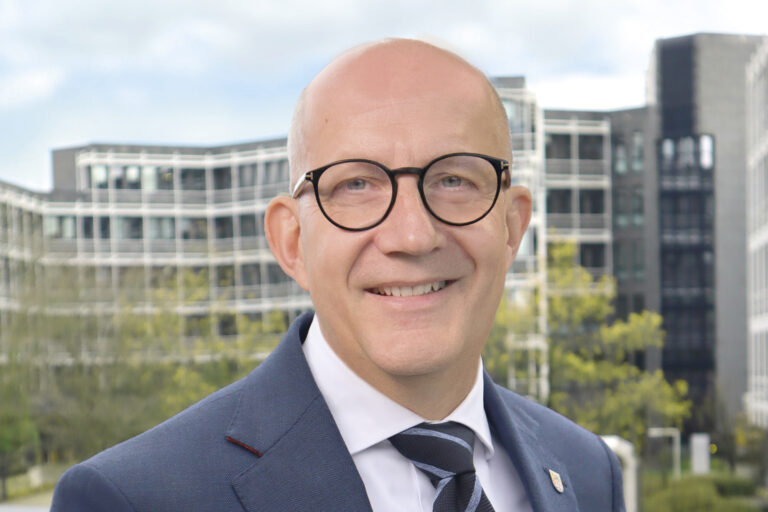 Ein Mann mit Glatze und runder Brille im Anzug lächelt vor modernen Bürogebäuden im Hintergrund.