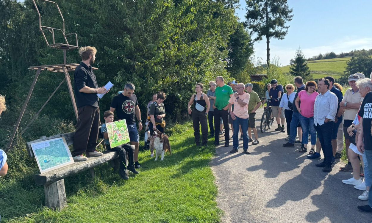 Gruppe von Menschen steht bei einer Führung an einem Feldweg; ein Mann erklärt etwas, während ein Kind eine selbstgemalte Karte zeigt.