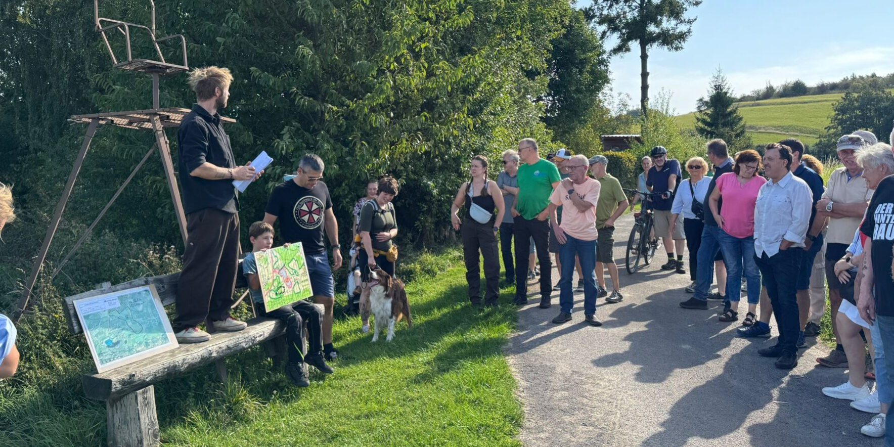Gruppe von Menschen steht bei einer Führung an einem Feldweg; ein Mann erklärt etwas, während ein Kind eine selbstgemalte Karte zeigt.