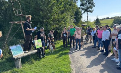 Gruppe von Menschen steht bei einer Führung an einem Feldweg; ein Mann erklärt etwas, während ein Kind eine selbstgemalte Karte zeigt.