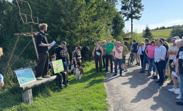 Gruppe von Menschen steht bei einer Führung an einem Feldweg; ein Mann erklärt etwas, während ein Kind eine selbstgemalte Karte zeigt.