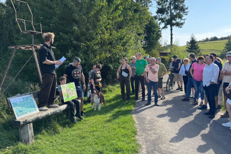 Gruppe von Menschen steht bei einer Führung an einem Feldweg; ein Mann erklärt etwas, während ein Kind eine selbstgemalte Karte zeigt.