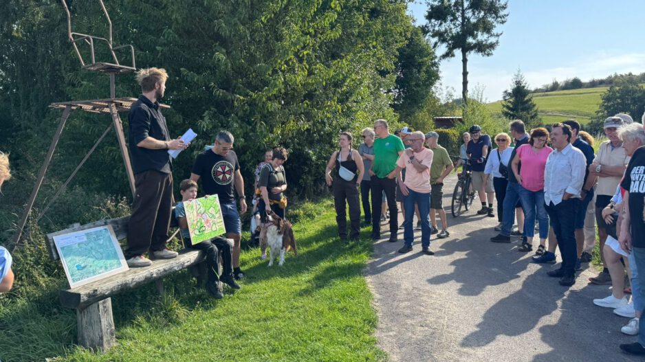 Gruppe von Menschen steht bei einer Führung an einem Feldweg; ein Mann erklärt etwas, während ein Kind eine selbstgemalte Karte zeigt.