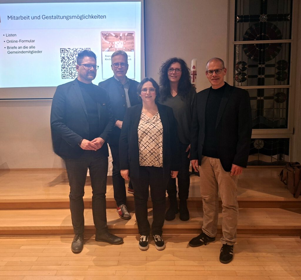 Fünf Personen stehen vor einer Präsentationsfolie in einem Veranstaltungsraum und blicken in die Kamera.