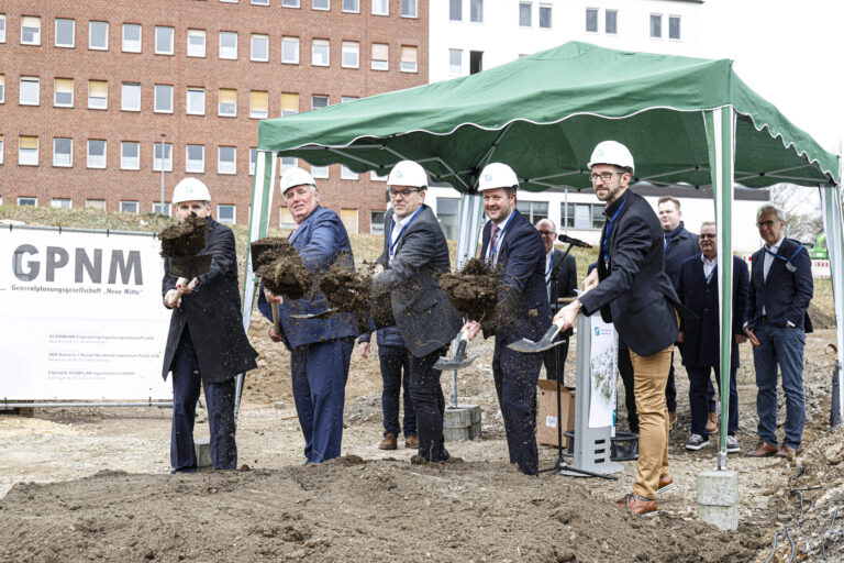 Mehrere Männer mit Schutzhelmen werfen bei einem symbolischen ersten Spatenstich Erde in die Luft.