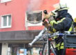 Feuerwehrleute löschen einen Brand in einem Gebäude; aus einem zerstörten Fenster steigt Rauch.