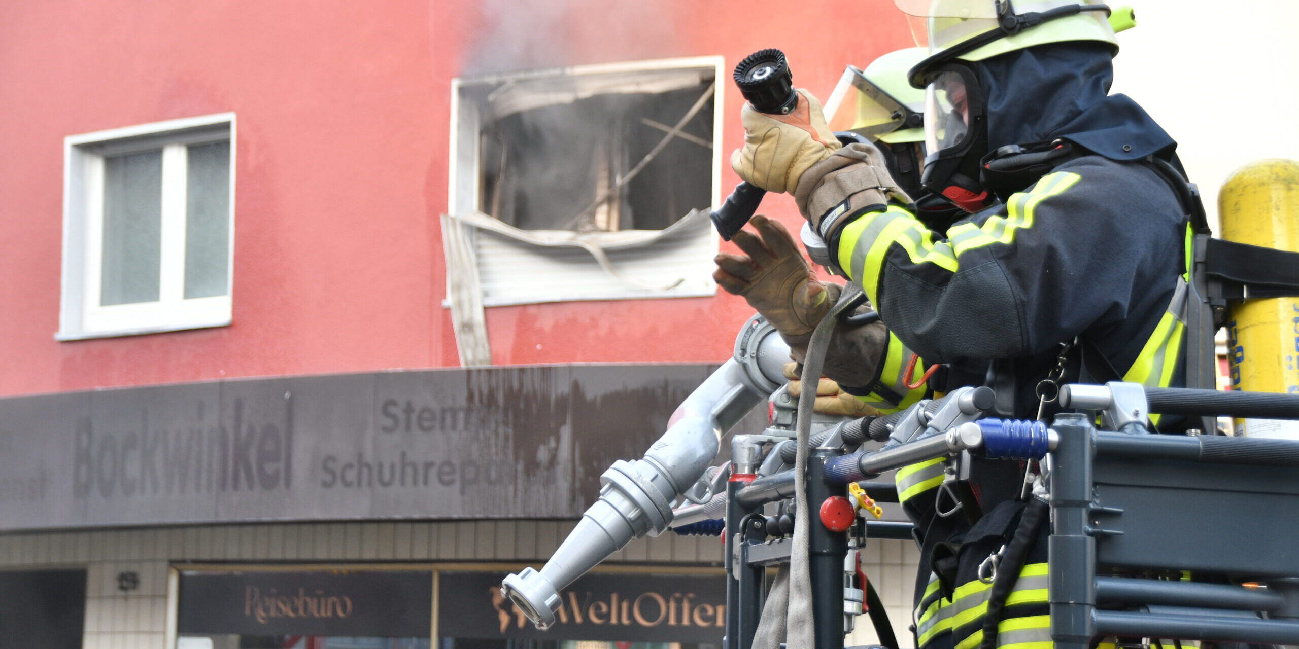 Feuerwehrleute löschen einen Brand in einem Gebäude; aus einem zerstörten Fenster steigt Rauch.