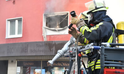 Feuerwehrleute löschen einen Brand in einem Gebäude; aus einem zerstörten Fenster steigt Rauch.