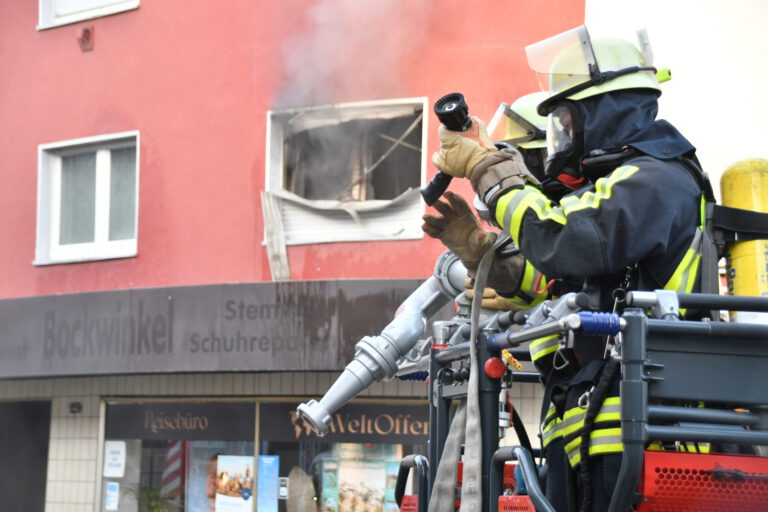 Feuerwehrleute löschen einen Brand in einem Gebäude; aus einem zerstörten Fenster steigt Rauch.