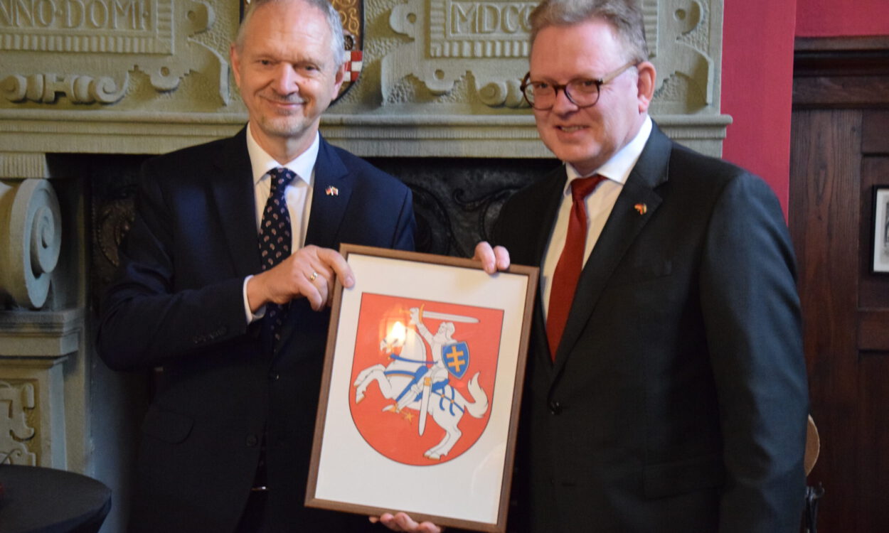 Zwei Männer in Anzügen halten gemeinsam ein gerahmtes Wappen mit einem Reiter auf einem Pferd vor einem Kamin.