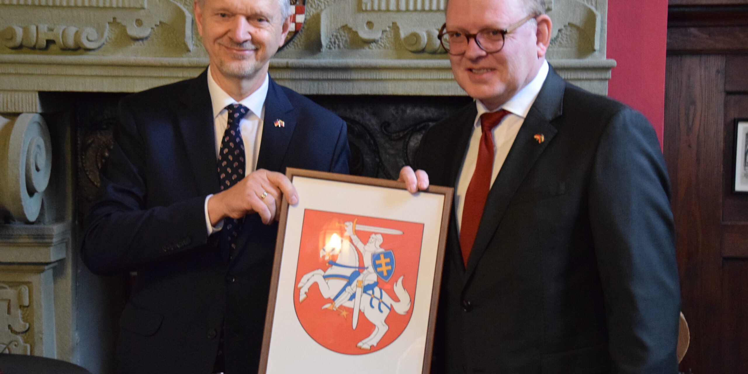 Zwei Männer in Anzügen halten gemeinsam ein gerahmtes Wappen mit einem Reiter auf einem Pferd vor einem Kamin.
