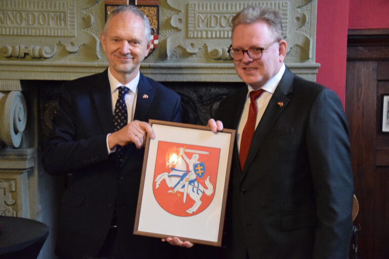 Zwei Männer in Anzügen halten gemeinsam ein gerahmtes Wappen mit einem Reiter auf einem Pferd vor einem Kamin.