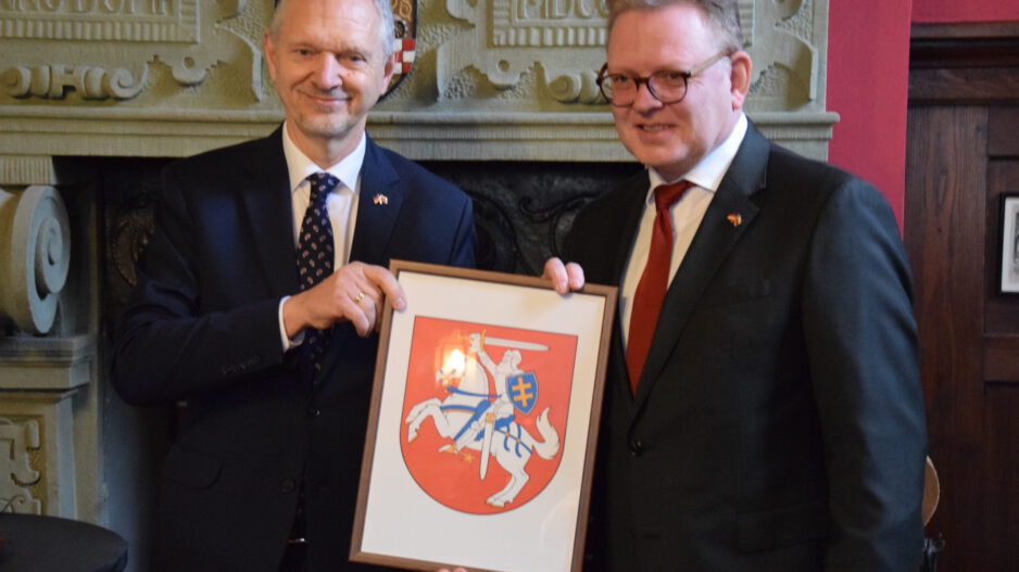 Zwei Männer in Anzügen halten gemeinsam ein gerahmtes Wappen mit einem Reiter auf einem Pferd vor einem Kamin.
