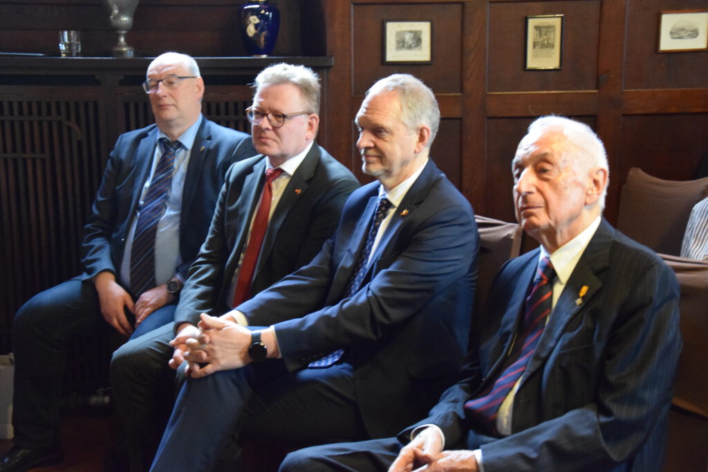 Vier Männer in Anzügen sitzen nebeneinander in einem holzvertäfelten Raum und hören aufmerksam zu.